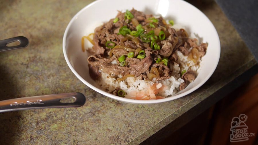 Finished and plated Japanese gyudonburi (beef rice bowl) sits on counter topped with sliced green onion and a side of pickled ginger waiting on a hungry dood to come and chow down. Looks delicious!