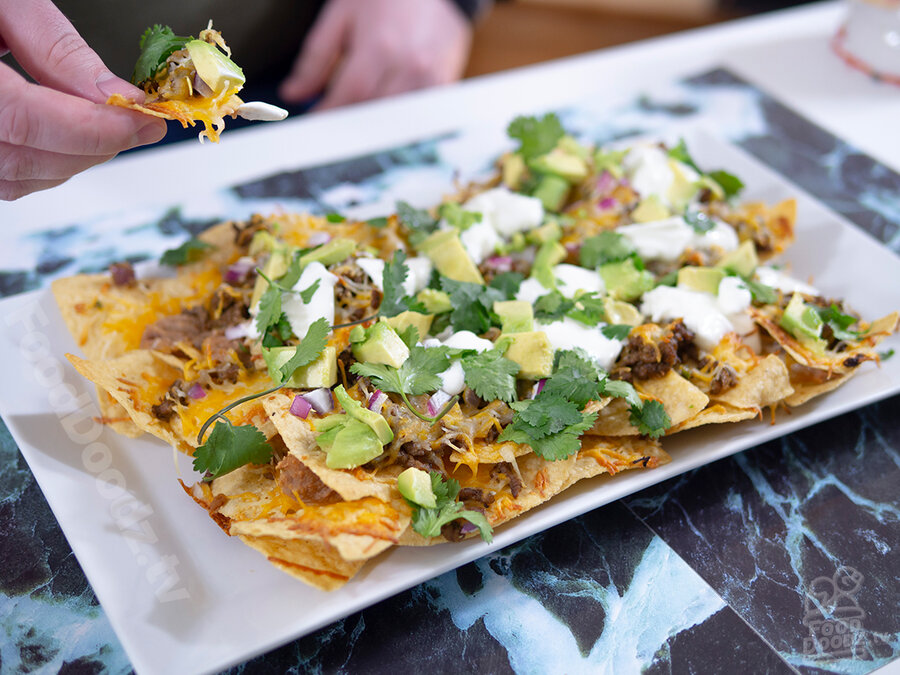 Gigantic rectangular plate of ultimate nachos sits on top of colorful background while hand holds a tasty bite above plate