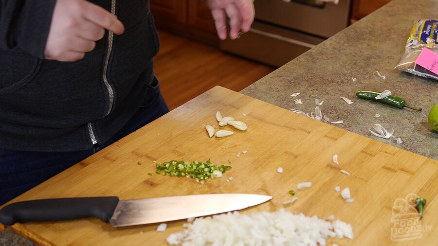 Peeled garlic gloves sit on cutting board next to chopped serrano pepper and onion