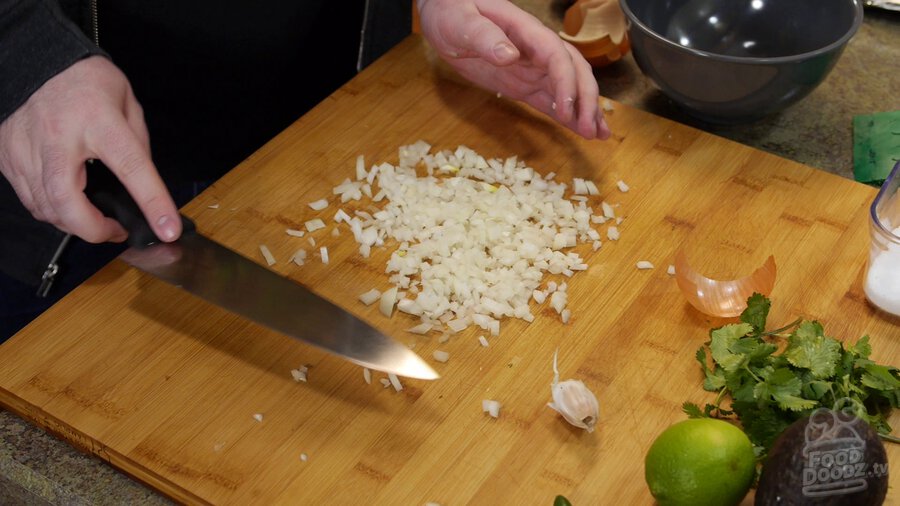 Chopped onion sits on wooden cutting board. Garlic, lime, avocado, and cilantro can be seen nearby.