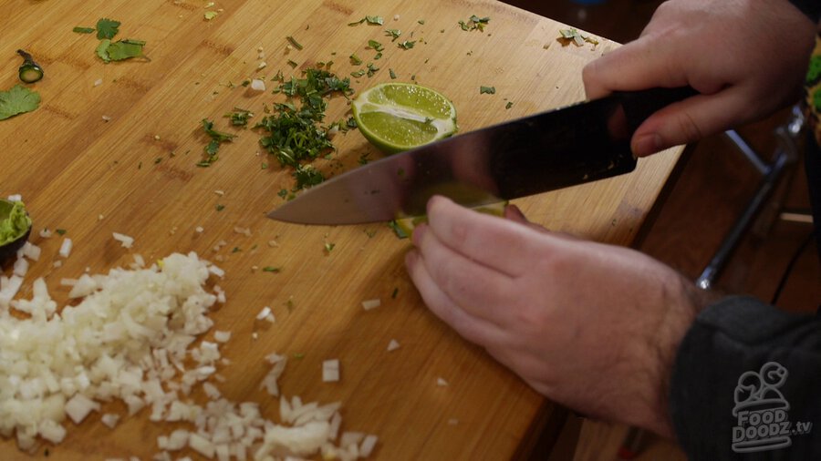 Lime is sliced with chef's knife on wooding cutting board to add lime juice to guacamole. Chopped onion and cilantro can be seen around.