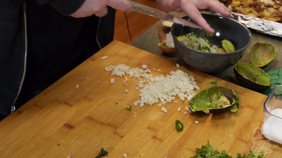 Using chef's knife serrano is scooped up and placed in bowl with avocado and onion.