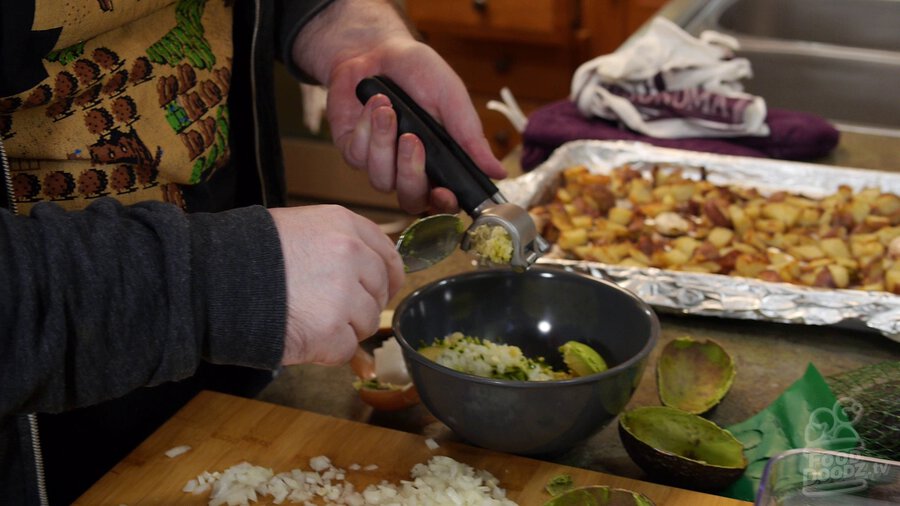 Using garlic press to mince garlic into bowl with avocado, onion, and serrano pepper.