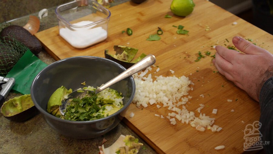 Finely chopping cilantro on wooding cutting board and adding it to bowl with avocado, onion, and serrano peppers.