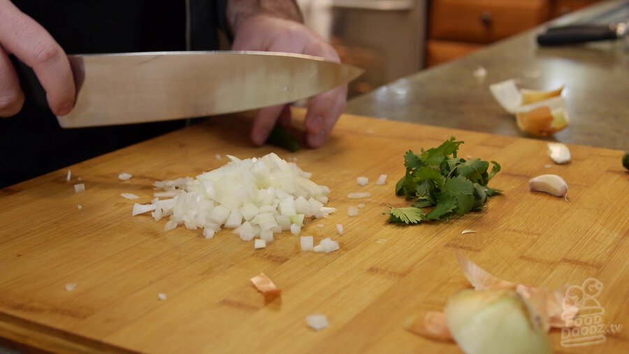 Onion sits chopped on cutting board next to a bunch of cilantro, while serrano pepper gets sliced
