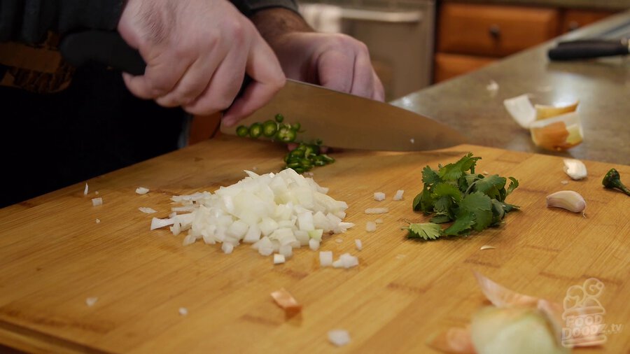 Chef's knife slicing up serrano pepper on cutting board. Chopped onion and a bunch of cilantro can be seen in the foreground.