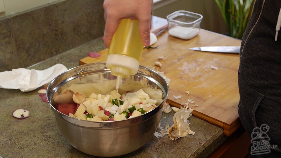 Using squirt bottle to add vegetable oil to large metal bowl filled with chopped/chunked potatoes, serrano peppers, and onions