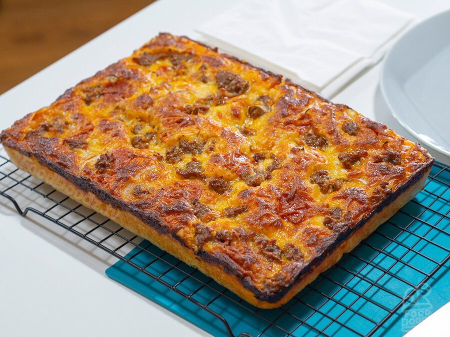 Absolutely beautiful golden brown sheet pan pizza resting on baking rack to cool. There's a nice caramelized cheese ring around the edge of the crust. The pie is topped with spicy Italian sausage, pepperoni, cheese blend, and onions. Yum!