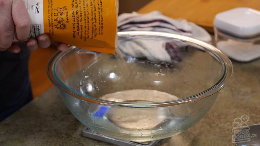 Yeast being poured out of bag into large glass bowl full of water and resting on digital scale