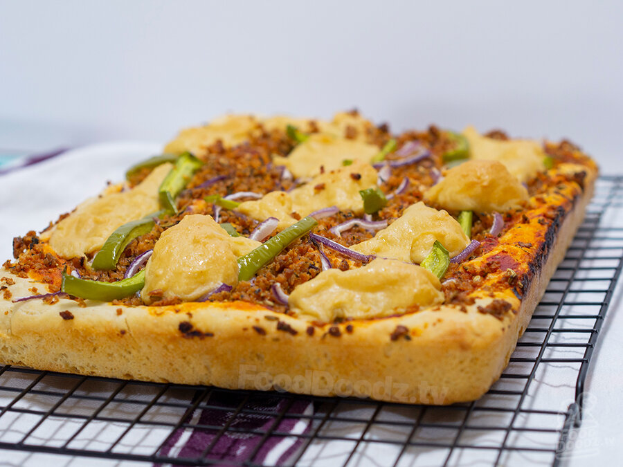 Golden brown and delicious (GBD) deep pan vegan pizza viewed from corner-on angle. Pie is covered in golden globs of vegan cashew cheese, homemade vegan Italian sausage, green peppers, and red onions