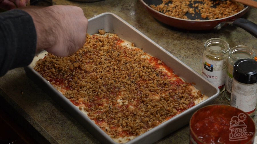 Sprinkling vegan textured vegetable protein (TVP) Italian sausage on top of pizza dough in sheet pan