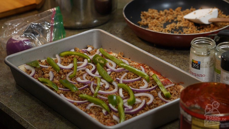 Slices of green peppers and red onion top pizza dough in sheet pan