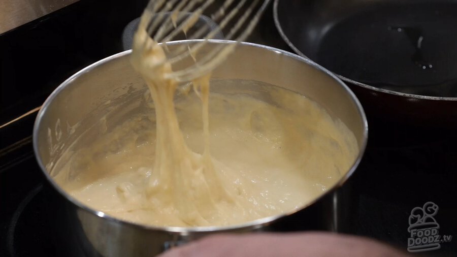 Whisk being pulled out of sauce pan to demonstrate stringy consistency of completed vegan cashew cheese recipe