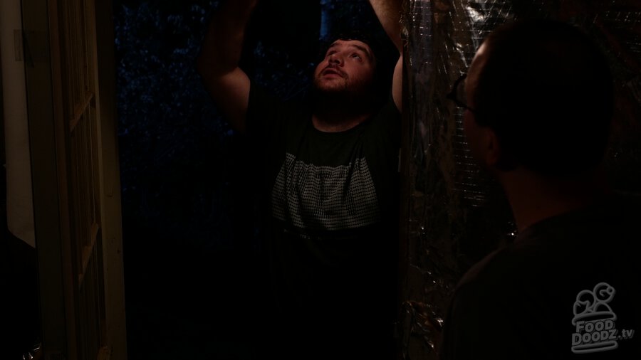 Adam standing in darkened doorway holding light above his head with his face illuminated. Austin stands nearby in darkness.