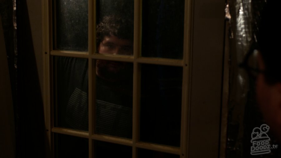 Adam standing in darkness knocking on window of door. He is barely illuminated. Austin's head is a dark blob across the right side of the screen.