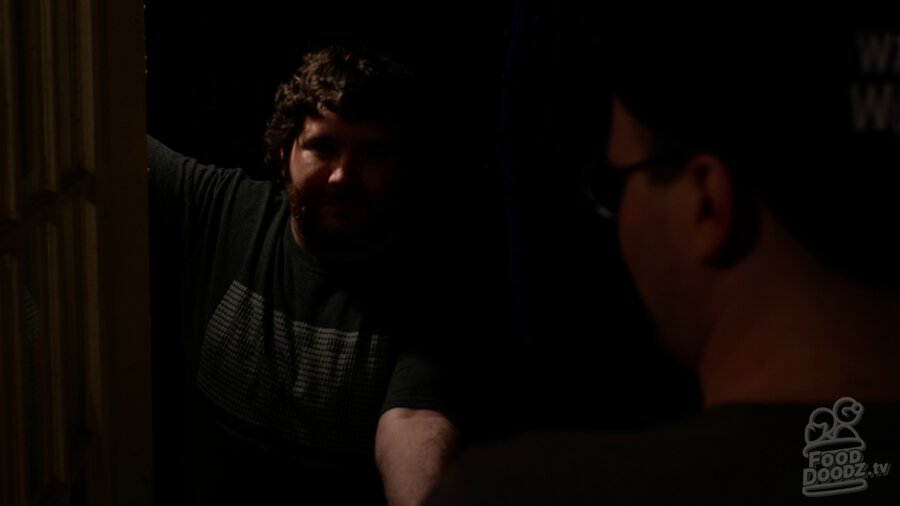 Adam standing in darkness through doorway. He is barely illuminated. He's handing Austin something. Austin's head and shoulders are a dark blob across the right third of screen.