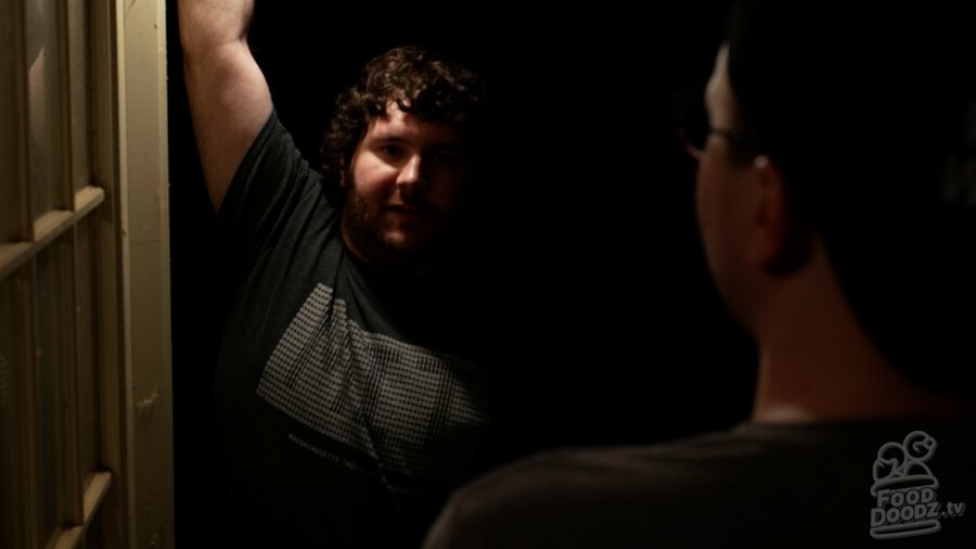 Adam standing in darkness through doorway. He is harshly illuminated from above on one side of his face. Austin's head and shoulders are a dark blob across the right third of screen.