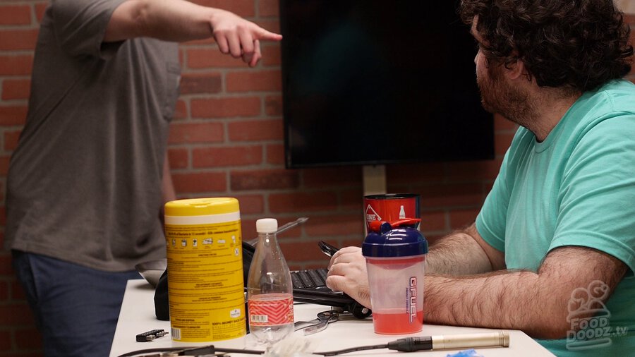 Adam sits at table while looking at Austin pointing at him from the left side. Austin's head is outside the top of the image frame.