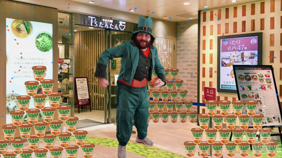 Adam dressed as leprechaun joyfully dancing in front of T's Tan Tan Ramen shop in Ueno Station in Tokyo with stacks of instant ramen surrounding him